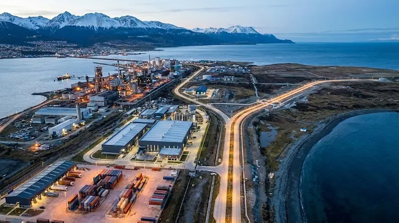 Vista aérea de un polo industrial y portuario en la costa de Tierra del Fuego, con rutas iluminadas, depósitos y montañas al fondo.