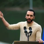 Nayib Bukele habla en un atril con micrófonos durante un anuncio educativo, con la bandera de El Salvador al fondo
