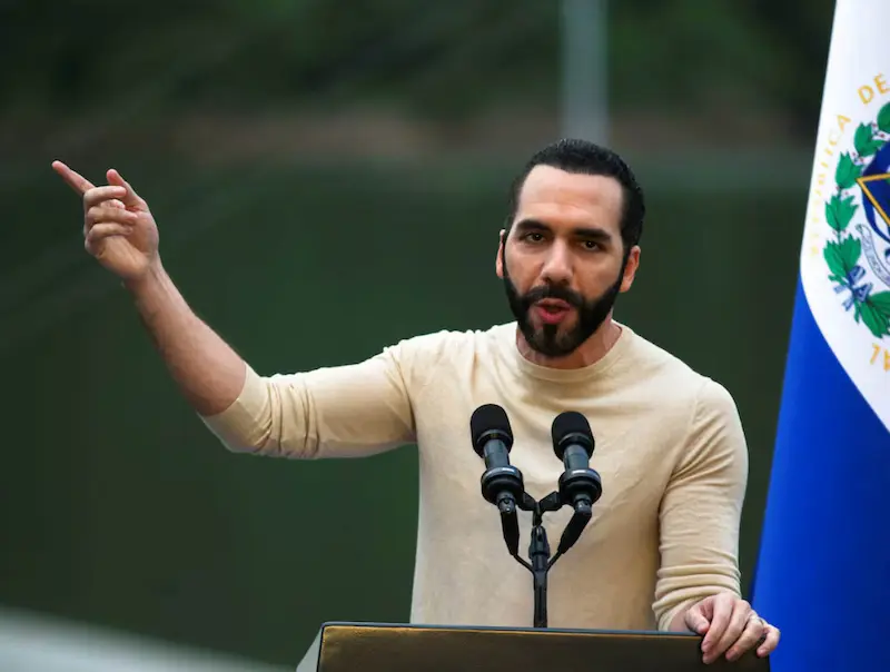 Nayib Bukele habla en un atril con micrófonos durante un anuncio educativo, con la bandera de El Salvador al fondo